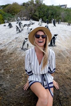 a woman in a hat and sunglasses sitting on the ground next to penguins with their mouths open