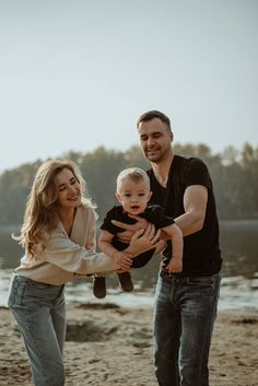 a man and woman holding a baby on the beach with trees in the back ground