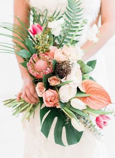 a bride holding a bouquet of flowers and greenery in her hands, with palm leaves on the side