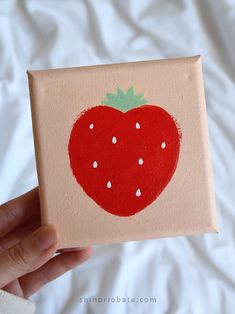 a hand holding up a small canvas with a strawberry painted on it's side