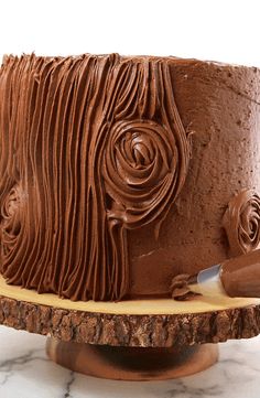 a close up of a chocolate cake with icing on a wooden stand and marble counter top