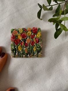 a hand holding a small square beaded brooch with flowers and leaves on it