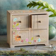 a wooden cabinet with flowers painted on the front and sides, sitting next to a green plant