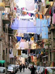 people walking down an alley way with clothes hanging from the buildings and cars parked on the street