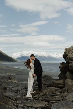 a bride and groom standing on rocks by the water with mountains in the background at their wedding