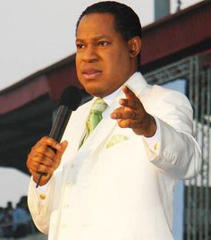 a man wearing a white suit and holding a microphone in his right hand while standing next to a stage