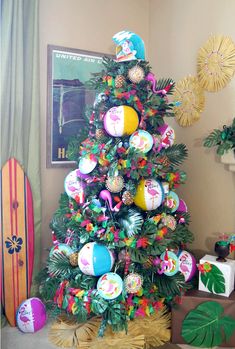 a colorful christmas tree decorated with beach balls and palm leaves, sits in front of a surfboard