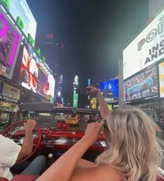 two people in a red convertible car driving through times square at night with neon signs and billboards