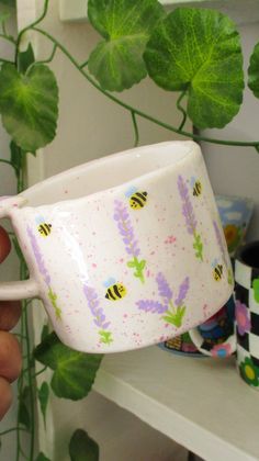 a hand holding a coffee cup with bees on it and purple flowers in the background