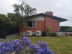 a red brick house with two air conditioners on the roof and purple flowers in front