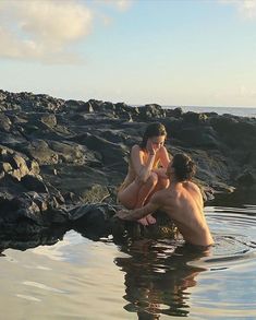 two people are sitting in the water near some rocks and one is on his cell phone