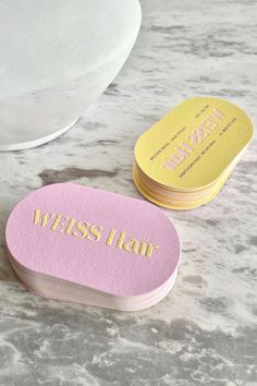 two pink and yellow business cards sitting on top of a marble counter next to a bowl