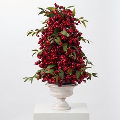 a vase filled with red berries and greenery on top of a white pedestal next to a wall