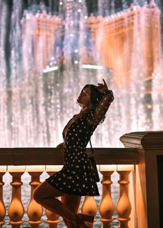 a woman in a black dress standing on a balcony next to a fountain at night