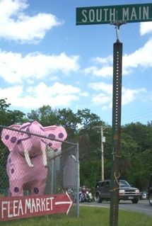 a pink elephant sign on the side of a road next to a street sign that says flea market