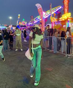 a woman in green jeans and white crop top walking through an amusement park at night