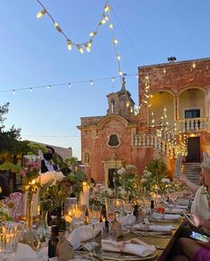 people are sitting at a long table in front of a building with lights strung over it