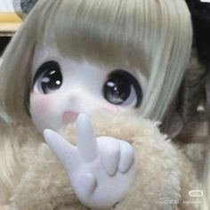 a close up of a stuffed animal with big eyes and blonde hair holding a peace sign