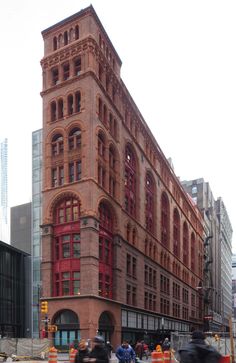 two people riding motorcycles down the street in front of a large building with red windows