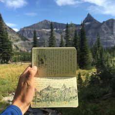 a person holding up an open book in front of some trees and mountains with writing on it