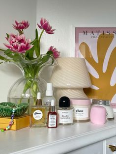 a white dresser topped with lots of bottles and vases filled with pink flowers next to a lamp