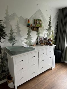 a white dresser sitting in front of a window next to a wall with trees on it