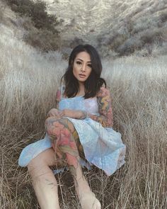 a woman sitting in the grass with her legs crossed and tattoos on her body, wearing a blue dress