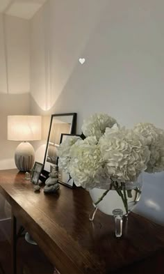 a vase with white flowers sitting on a wooden table next to a lamp and pictures