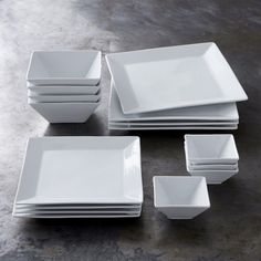 a set of white square plates and bowls on a gray table top with one empty plate in the middle