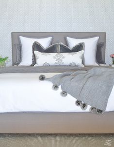 a bed with white and gray pillows and blankets on top of it next to flowers