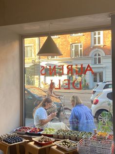 two people sitting at a table in front of a window with fruit and vegetables on it