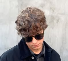 a young man with curly hair and sunglasses looking at the camera while standing in front of a concrete wall