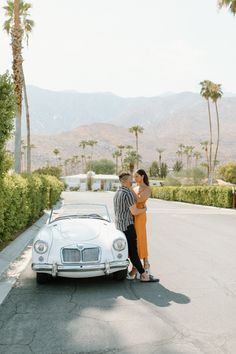 a man and woman standing in front of a car on the road with palm trees