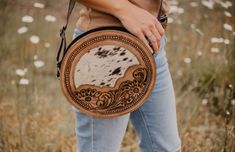 A boho-inspired canteen-style bag with an adjustable strap, featuring tooled floral leather on the front and a cowhide accent in the center, combines rustic charm with artistic flair. This unique blend of materials and design elements creates a trendy and eye-catching accessory, perfect for adding a bohemian touch to your everyday style. Round Western Purse, Custom Leather Work, Western Bag, Western Purses, Cute Country Outfits, Cowhide Bag, Hat Ideas, Cute N Country, Leather Work