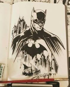 a drawing of batman in black and white on a book with marker pens next to it