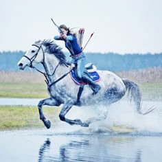 a woman riding on the back of a white horse across a body of water with trees in the background