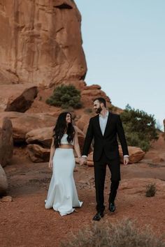 a man and woman holding hands walking through the desert with large rocks in the background