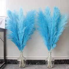 two clear vases with blue feathers in them on a marble floor next to a white wall