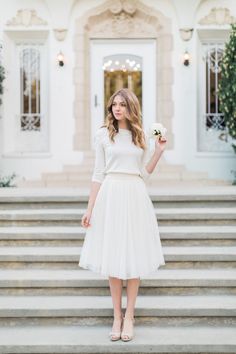 a woman in a white dress is standing on some steps holding a flower and looking at the camera