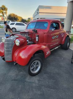 an old red truck parked in a parking lot next to a man working on the hood