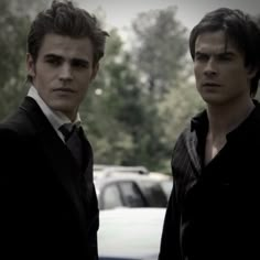 two young men standing next to each other in front of a car on the street