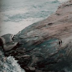 two people standing on the edge of a cliff by the ocean