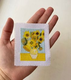 a hand holding a small cross stitch picture with sunflowers in a vase