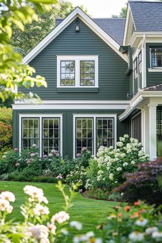 a green house with white trim and flowers in the front yard, surrounded by greenery