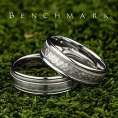 two silver wedding bands sitting on top of green grass with the words ben ch mark written