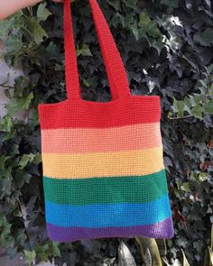 a hand holding a multicolored crocheted bag in front of a bush