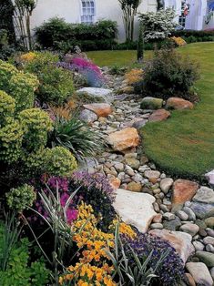 a garden with rocks and flowers in front of a white house on the side of a hill