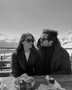 a man and woman sitting at a table in front of snow covered mountains with food on it