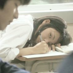 a woman is writing in a notebook while sitting at a desk with her head on the table