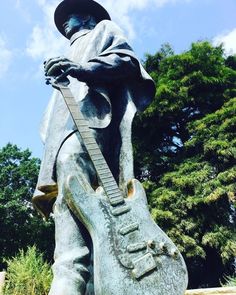 a statue of a man with a guitar in his hand and trees behind him on a sunny day